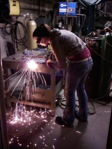 April welding at a workbench, sparks across the shop floor.